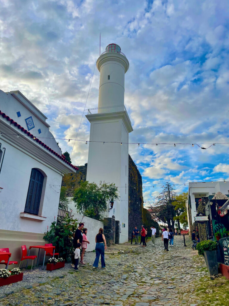 The iconic Colonia del Sacramento lighthouse standing tall against a blue sky, with remnants of the old convent at its base
