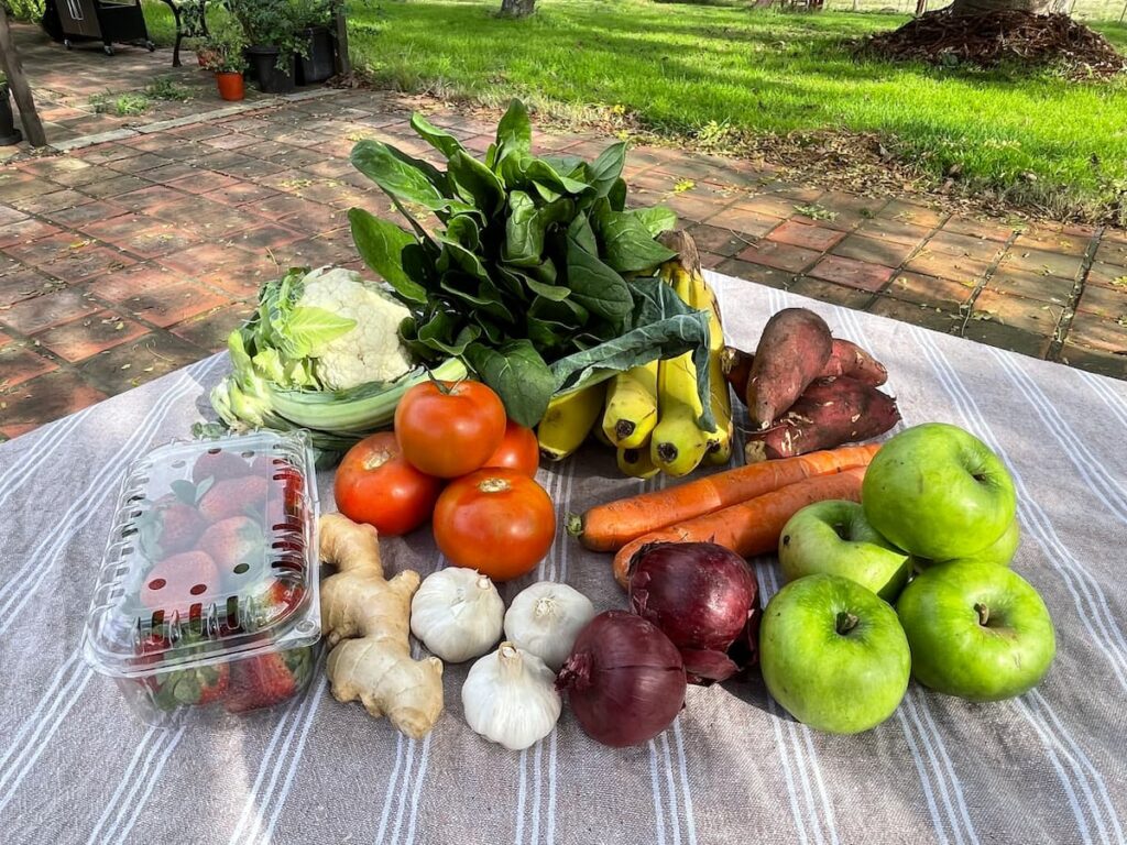 Fresh produce display in a rural Uruguay setting including strawberries, tomatoes, spinach, bananas, apples, garlic, ginger, red onions, cauliflower, and sweet potatoes on a rustic outdoor table, food quality in uruguay