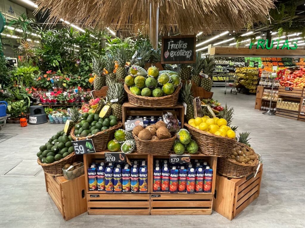 Fruit display in a Uruguayan supermarket showcasing a variety of exotic fruits including papaya, coconut, avocados, pineapple, and kiwi arranged in wooden crates under a tiki hut, food quality in uruguay